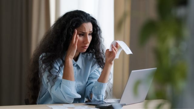 Mulher branca de cabelos escuros longos, sentada à mesa segurando papel com notebook aberto à frente