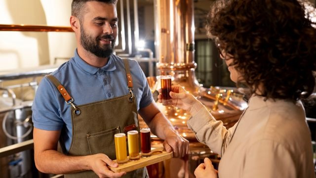 Em ambiente industrial, homem de avental segura bandeja segura copos de bebida enquanto mulher de cabelo cacheado segura um copo