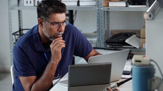 Homem de óculos sentado à mesa usando notebook e tablet em um ambiente de almoxarifado