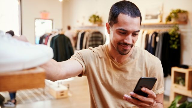 Homem utilizando celular em loja de roupas com araras e produtos ao fundo