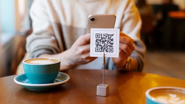 Pessoa apontando o celular para QR code em suporte sobre mesa de madeira com xícaras de café e pires azuis