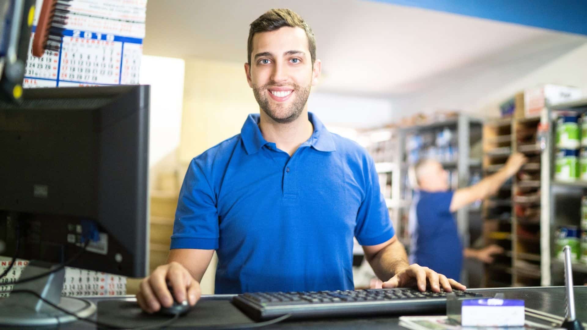 Pessoa em um balcão de loja usando o computador e teclado, ao fundo prateleiras de produtos e estoque.