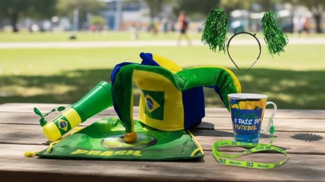 Corneta, mochila, chapéu, caneca e cordão nas cores do Brasil dispostos sobre mesa em ambiente de parque.