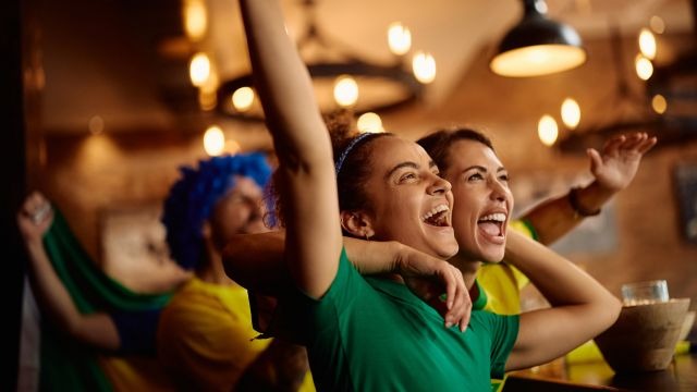 Duas mulheres vestidas nas cores do Brasil comemorando em um bar com outras pessoas ao fundo.