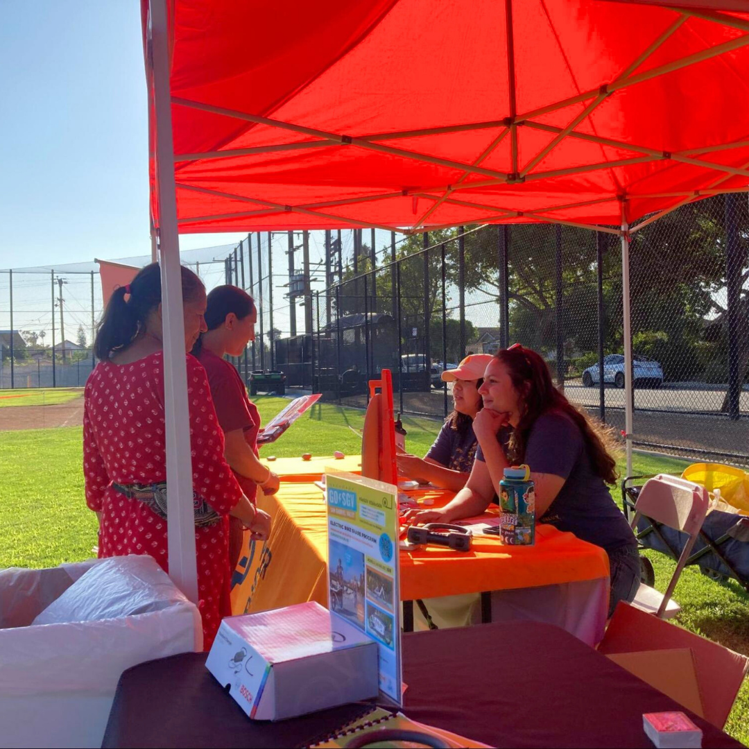 People speaking with staff at a booth under a red tent at a community park event.