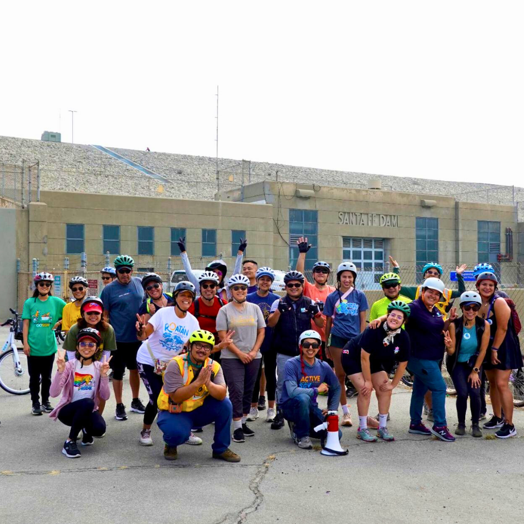 Group of cyclists wearing helmets posing for a photo in front of Santa Fe Dam during a bike ride event.