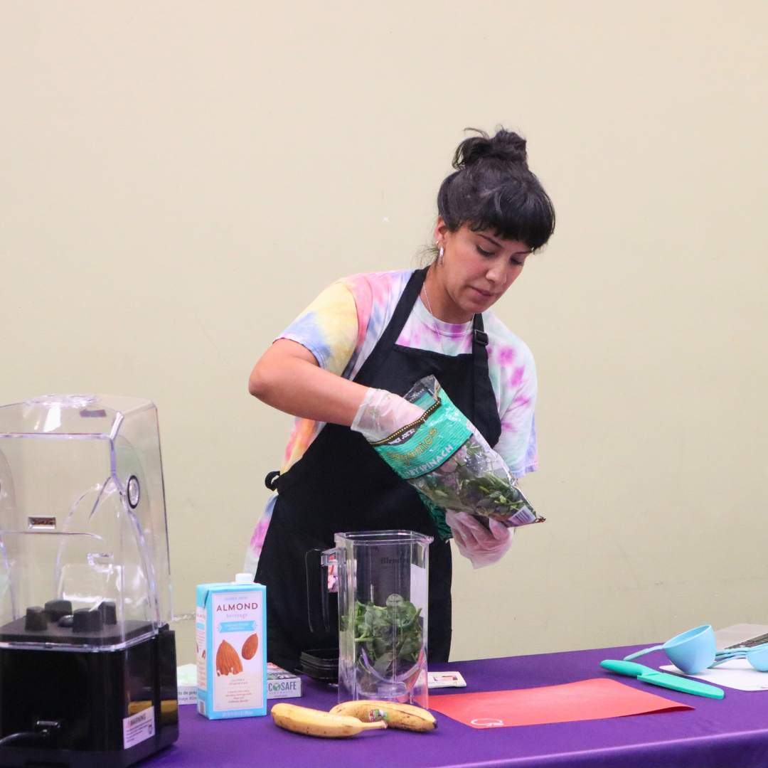 Woman in a tie-dye shirt and apron preparing a smoothie with spinach, bananas, and almond milk on a table with a blender.