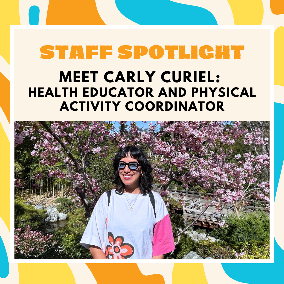 Carly Curiel stands smiling in front of blooming pink cherry blossoms, wearing sunglasses and a white shirt with a large flower graphic.