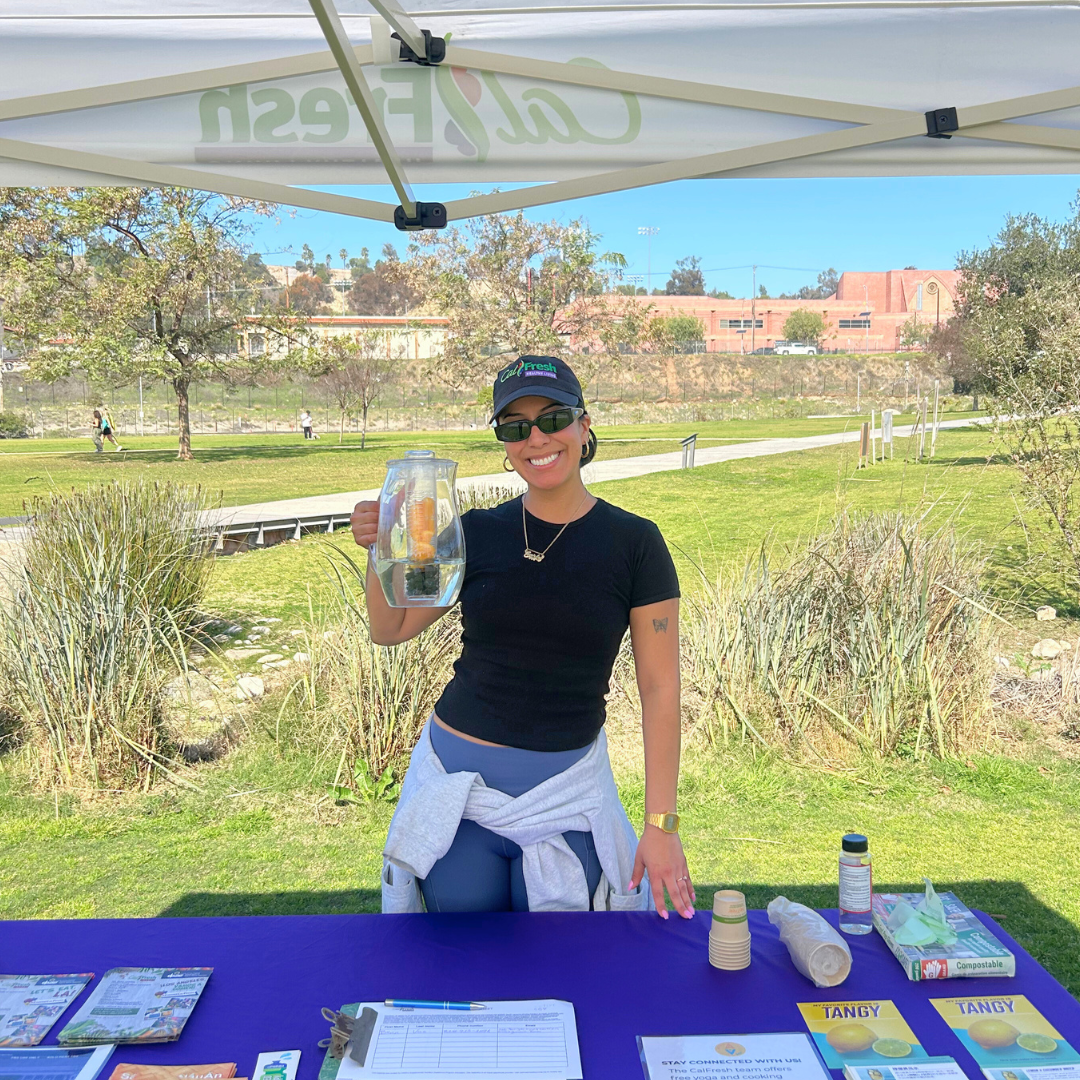 Carly stands at an outdoor CalFresh Healthy Living table holding a pitcher of fruit-infused water, smiling at the camera.