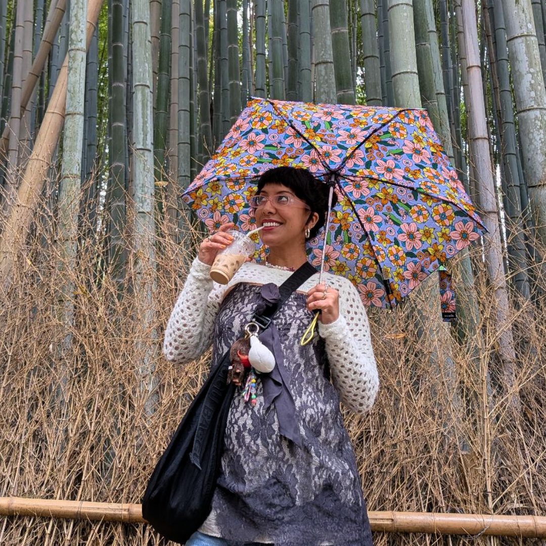 Carly stands in front of tall bamboo holding an iced drink and a colorful floral umbrella, smiling as she looks off to the side.