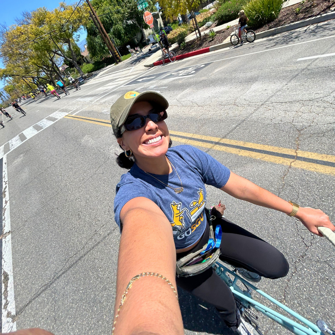 Carly smiles widely while riding her bike down a sunlit street during an open streets event, wearing a GoSGV t-shirt and baseball cap.