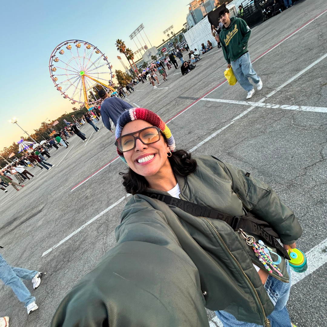Carly takes a selfie at a fairground, smiling with a Ferris wheel and festivalgoers in the background.