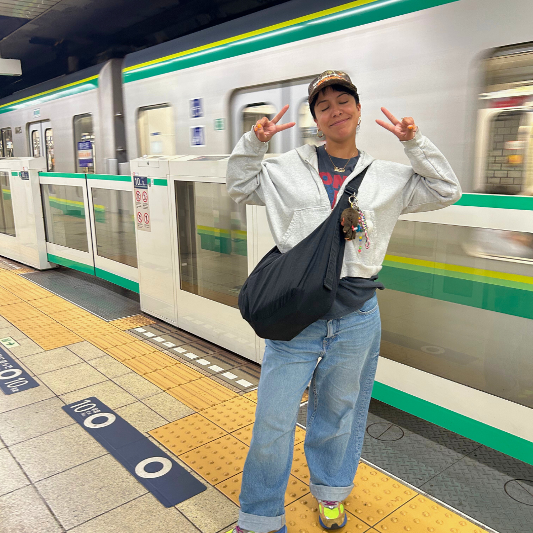 Carly holding up a peace sign on a train platform, wearing loose jeans, sneakers, and a baseball cap.