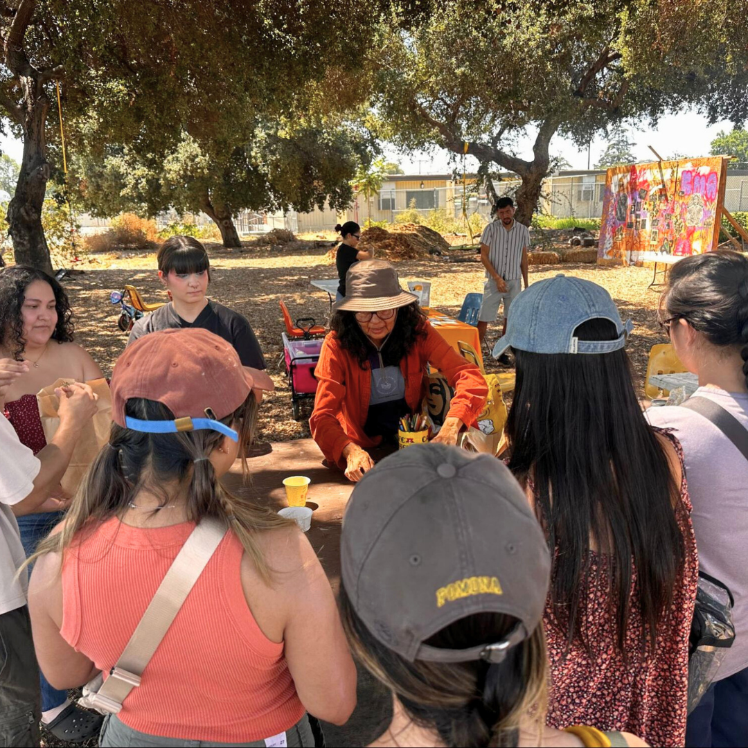 Community members gather around a table outdoors for a hands-on workshop led by an instructor in a hat and orange jacket.