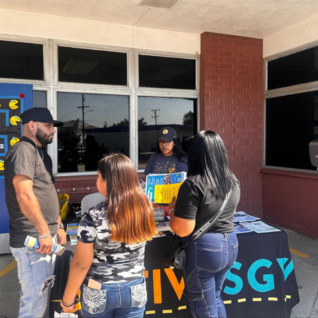An ActiveSGV staff member speaks with community members at an outreach table covered with flyers and resources.