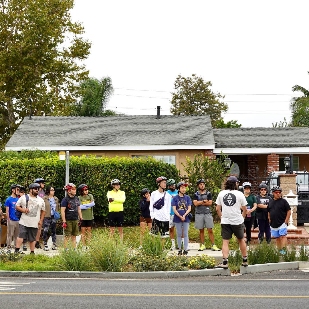A large group of cyclists stand together on a residential street listening to instructions.