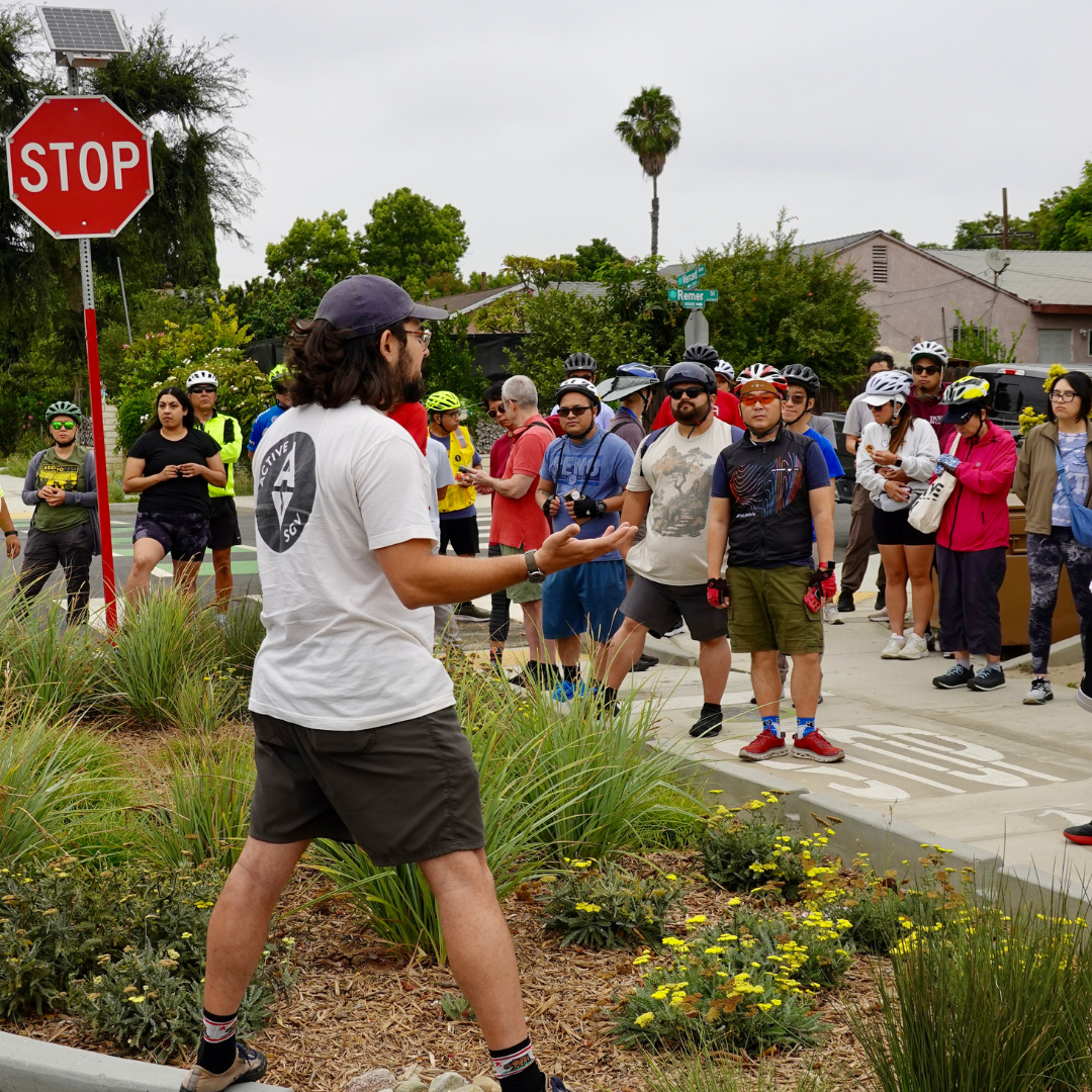 An ActiveSGV ride leader in a white shirt addresses a group of cyclists near a crosswalk and stop sign.