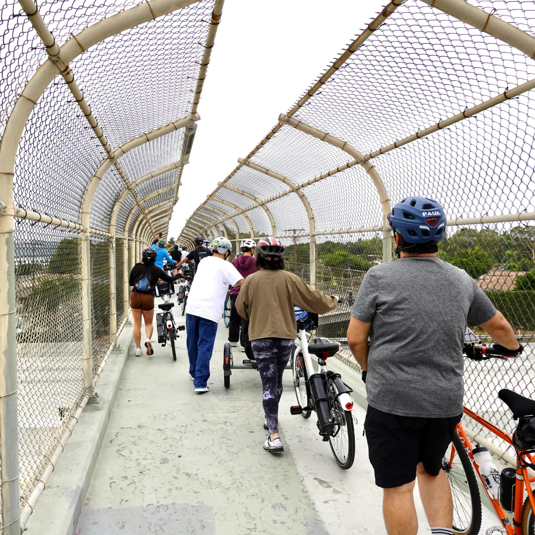 Cyclists walk their bikes across a fenced pedestrian bridge.