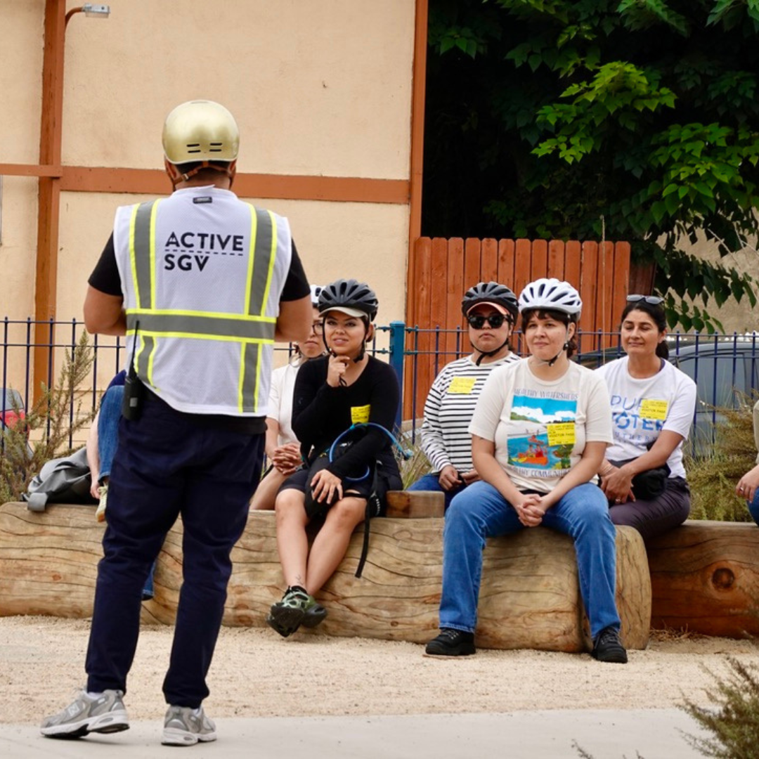 An ActiveSGV staff member wearing a reflective vest speaks to a seated group of cyclists in a park.