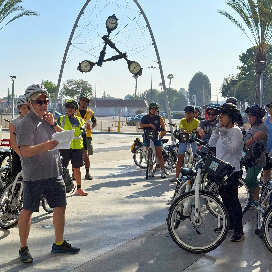 A group of cyclists gather at the El Monte Transit Center under the “Time” clock sculpture, listening to a speaker holding notes.