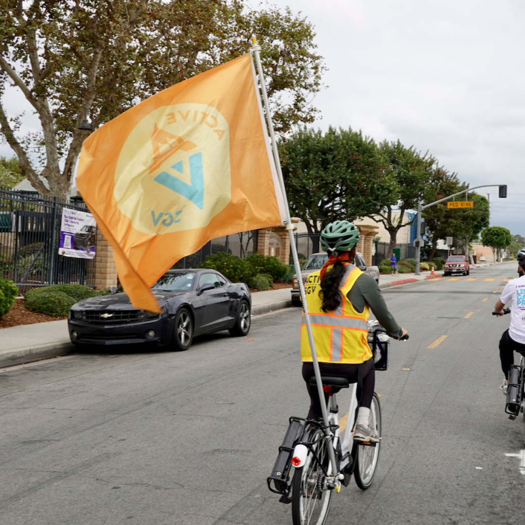 A cyclist rides down a city street carrying a large ActiveSGV flag attached to their bike.