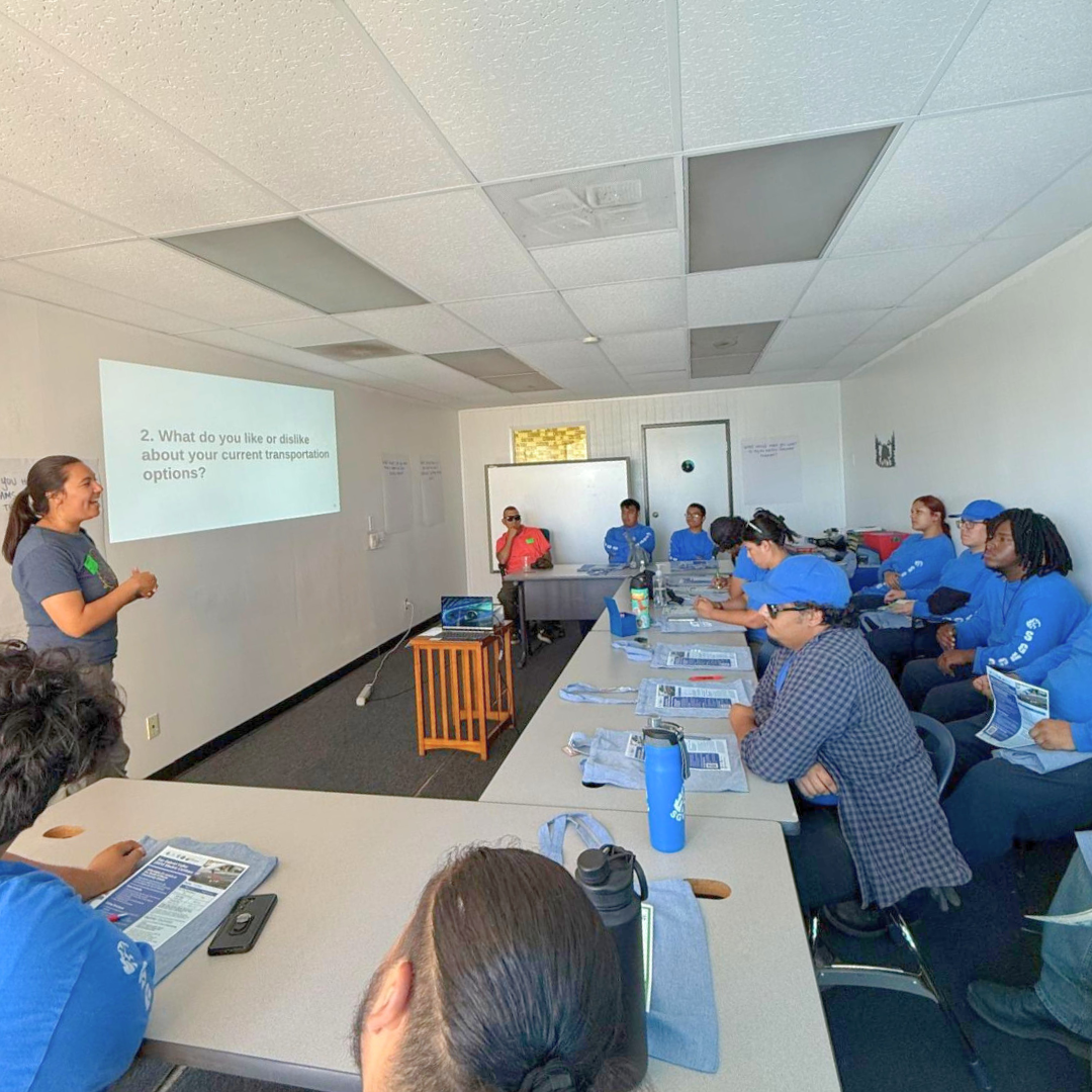 A facilitator leads a workshop as a group of young adults in blue shirts and hats listen and take notes, with a slide projected on the wall.