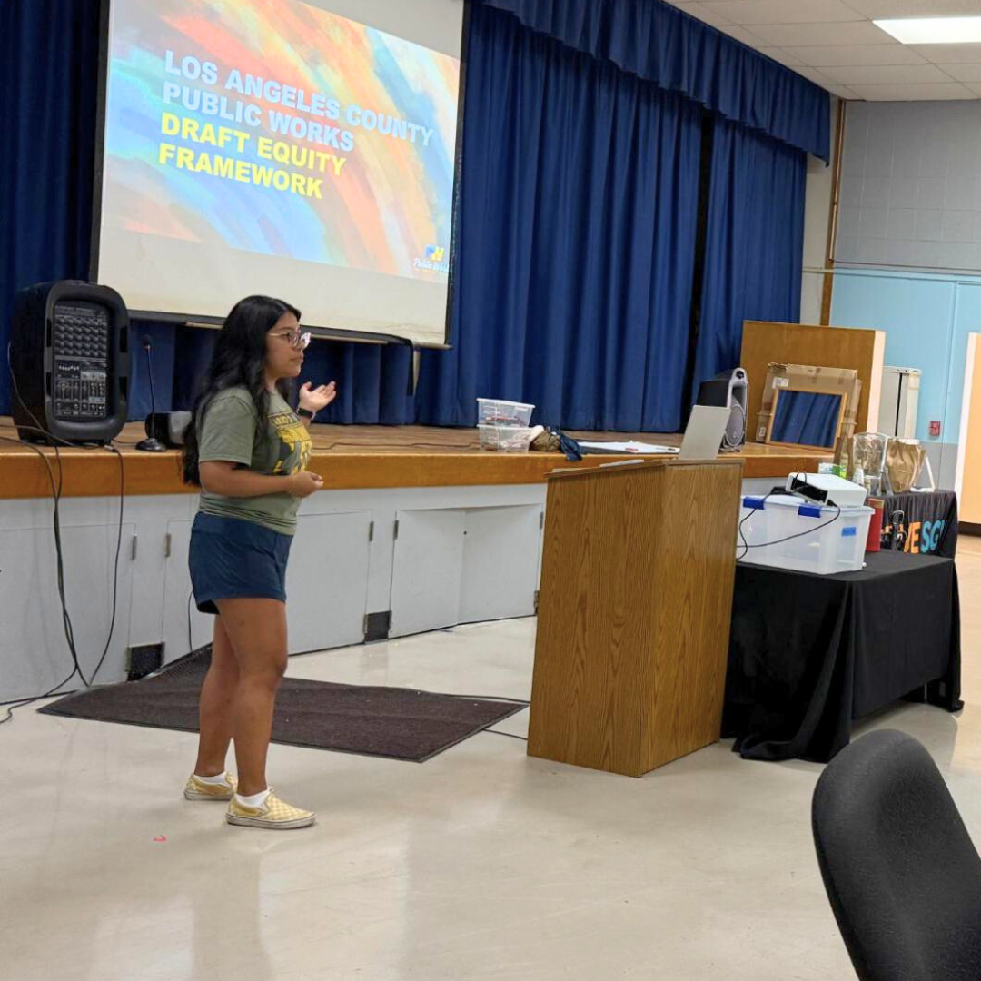 A woman presents on stage in front of a large screen displaying “Los Angeles County Public Works Draft Equity Framework.”