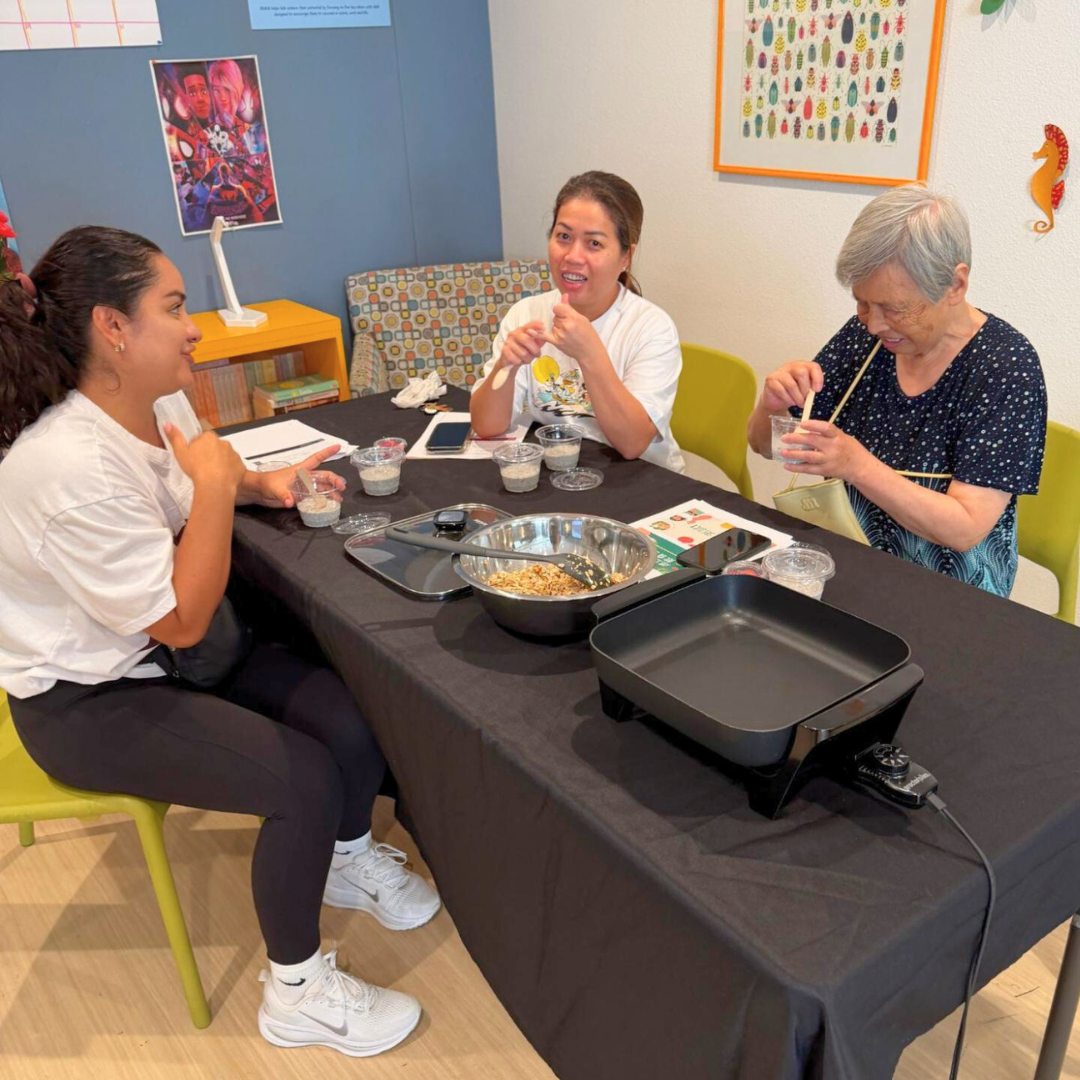 Three women sit around a table preparing food samples during a nutrition workshop, with mixing bowls and cups in front of them.