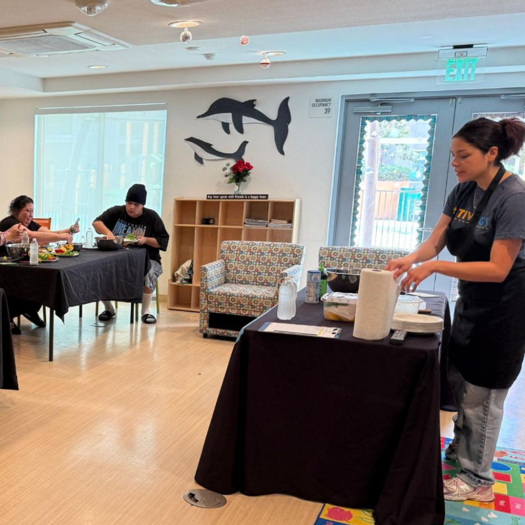 A facilitator stands at a table demonstrating a recipe while participants seated at nearby tables follow along.