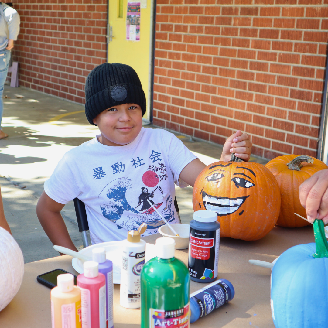 A child smiles while painting a pumpkin with bright colors at a community table.