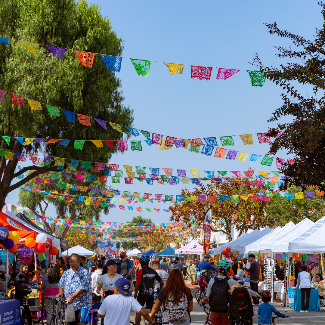 A busy street lined with vendor tents and colorful papel picado banners overhead as people walk, bike, and explore.