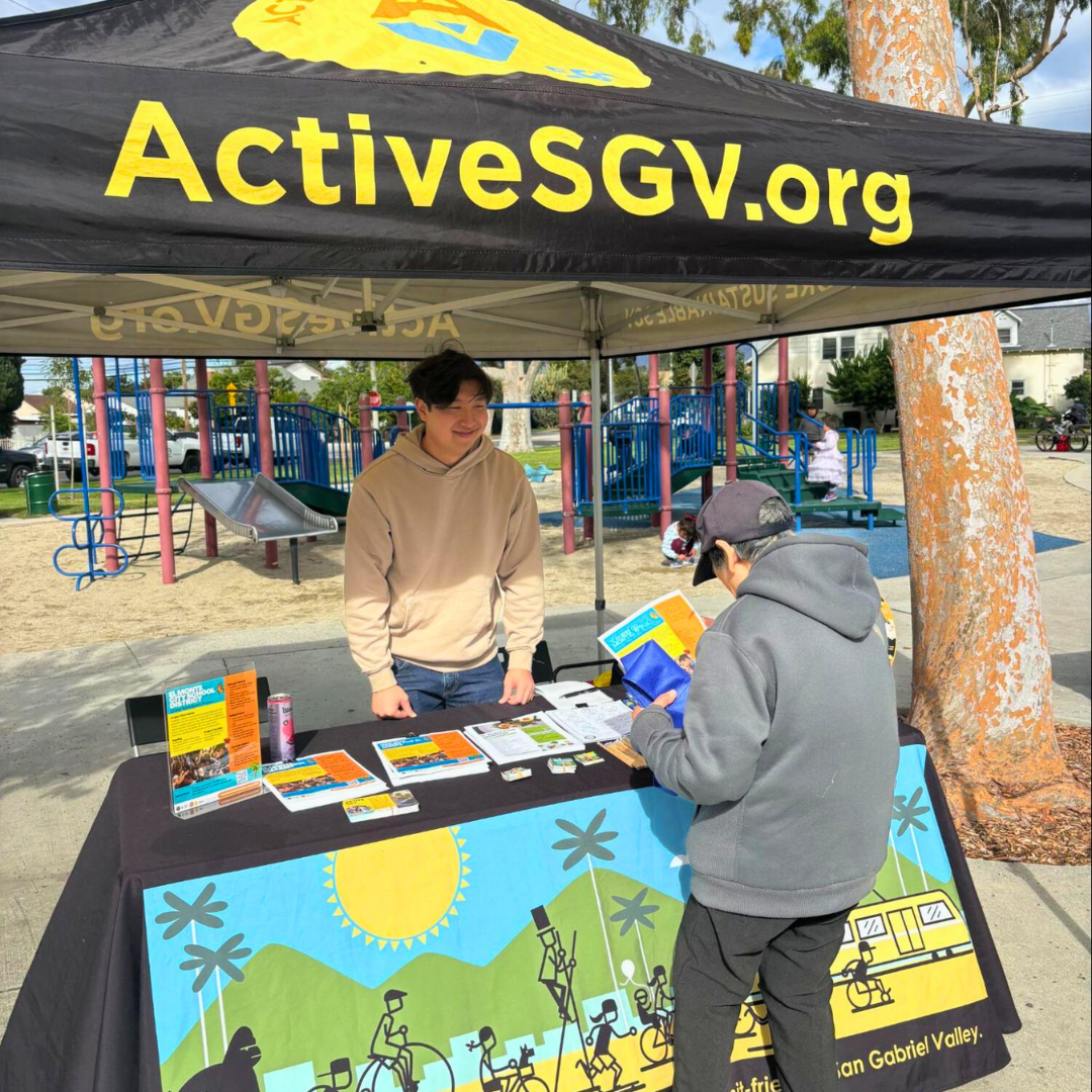 An ActiveSGV staff member speaks with a community member at an outdoor table covered with brochures in front of a playground.