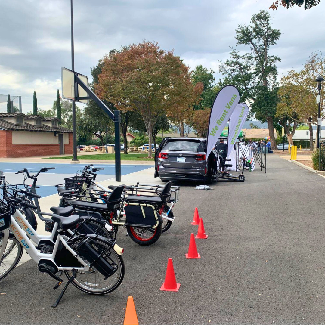 Several e-bikes and a van are set up near a basketball court with cones arranged for a demo course.