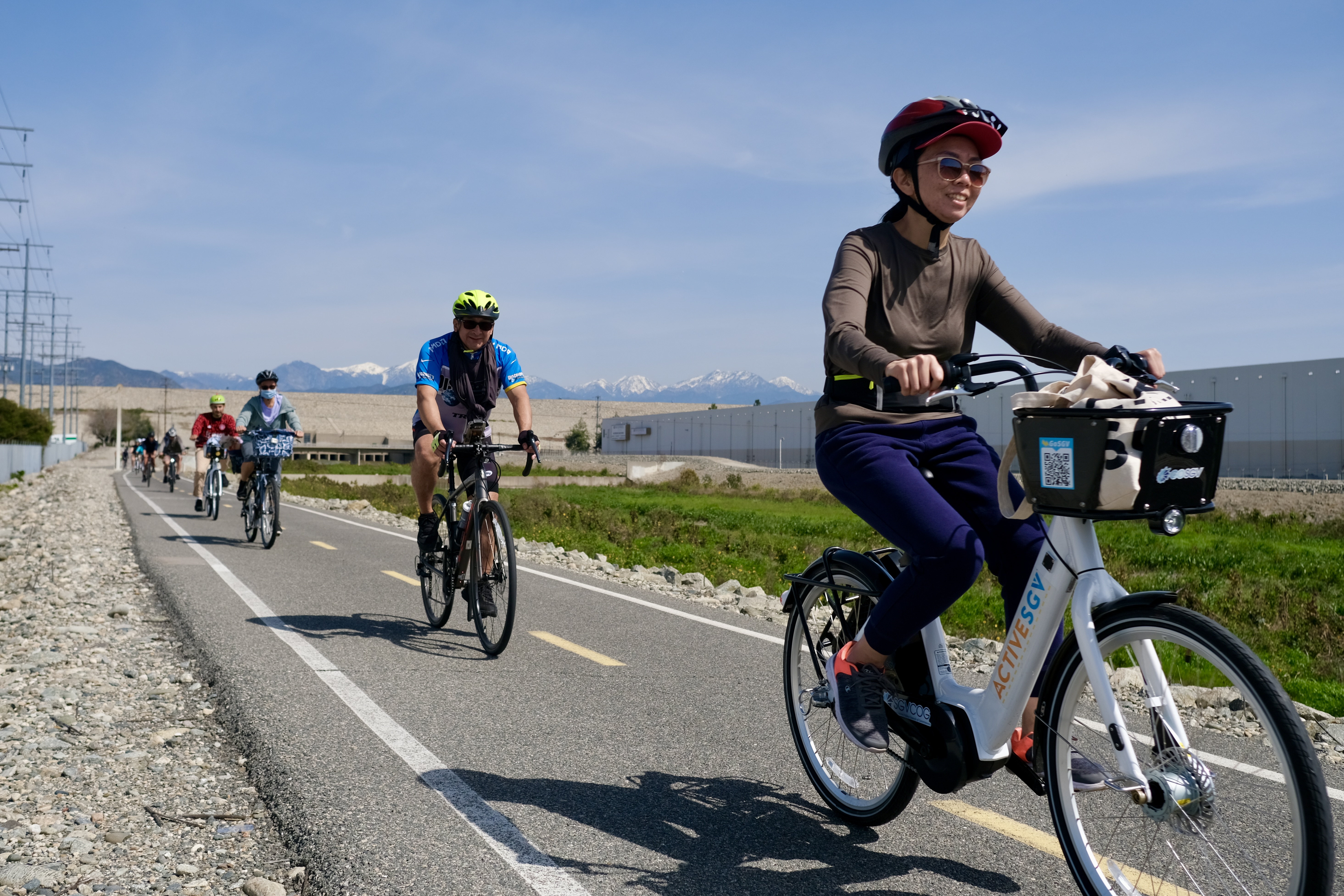 sunny outdoor scene showing a group of people riding bicycles on a paved, multi-use path. In the foreground, a smiling woman wears a helmet and rides a white electric bike with a front basket labeled "ActiveSGV." Behind her, a man in a blue jersey rides a road bike, followed by several other cyclists in the distance. To the left, high-voltage power lines run parallel to the path, and in the background, snow-capped mountains rise behind a row of industrial buildings.