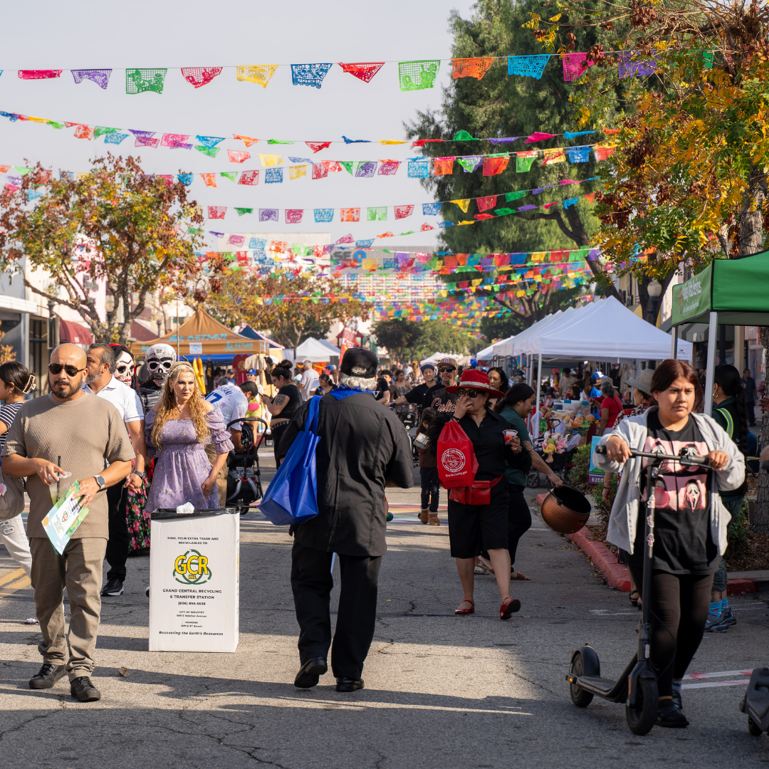 A crowded open street event with colorful papel picado overhead. People walk, shop, and explore vendor booths.