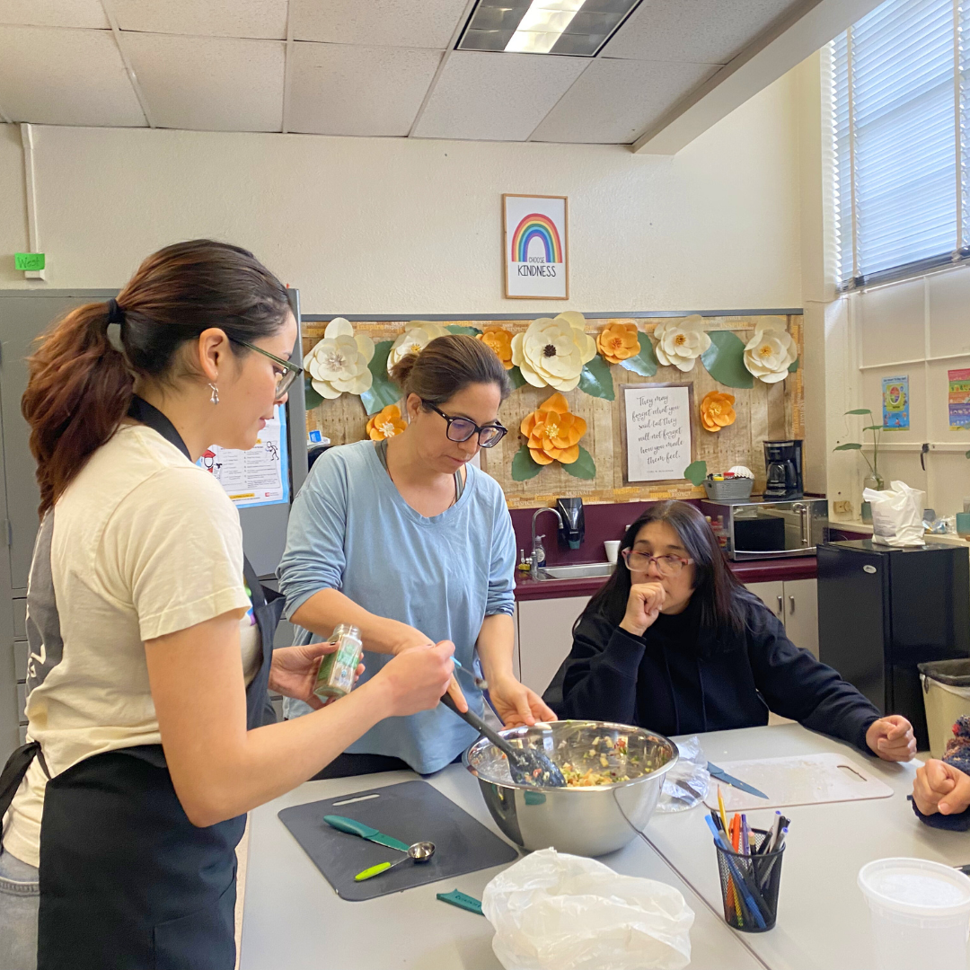 Three people gather around a table in a school kitchen, mixing and seasoning a large bowl of chopped vegetables, with paper flowers and a “Choose Kindness” sign on the wall.