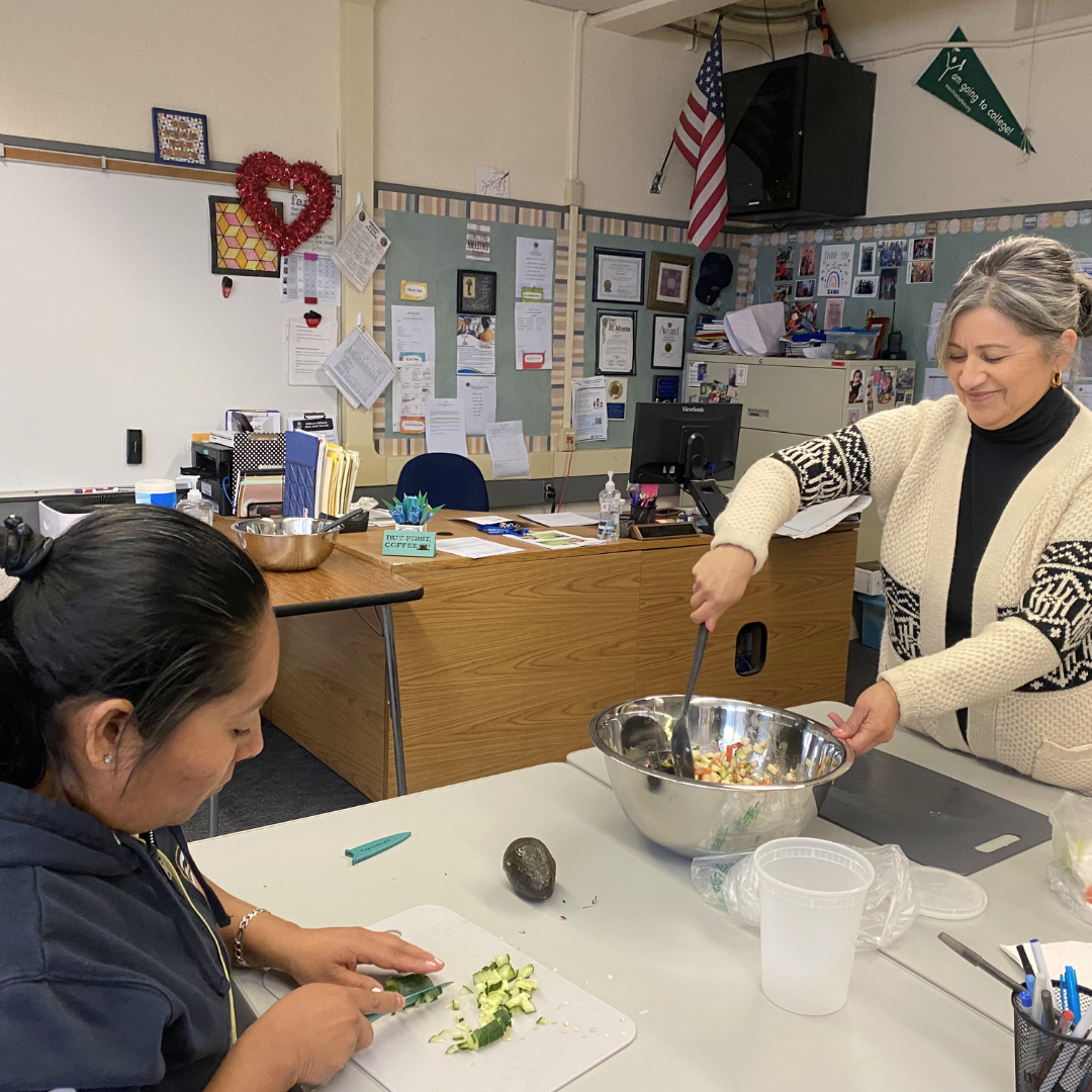 In a classroom, one participant chops cucumber on a cutting board while another mixes a large bowl of chopped vegetables at a table with an avocado and utensils.
