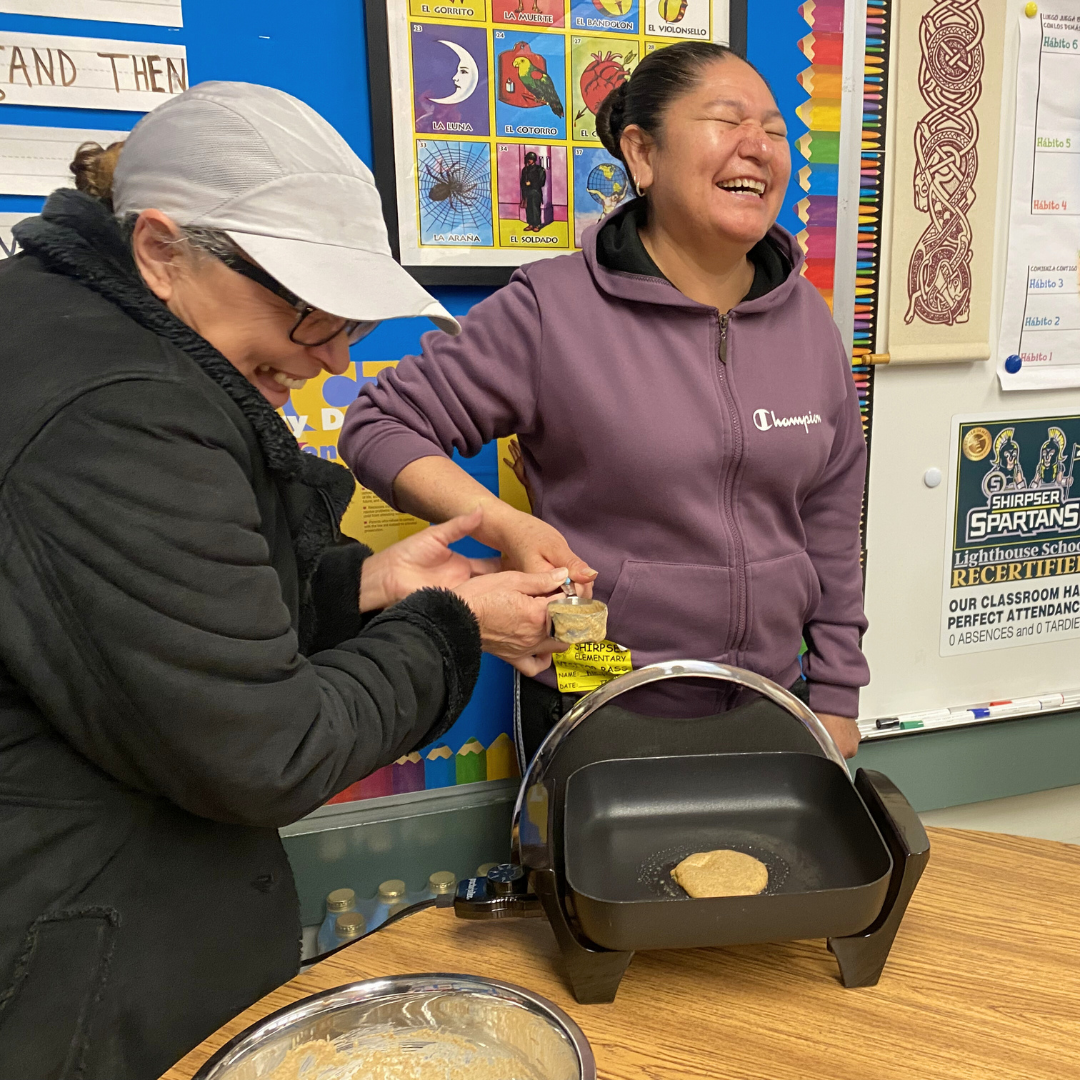 Two participants laugh while working next to an electric griddle cooking a pancake, with classroom posters in the background.