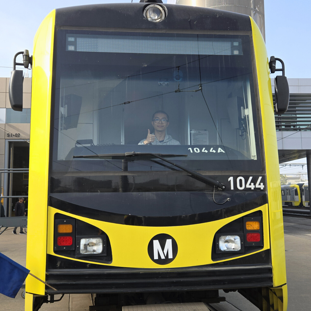 Front view of a yellow-and-black light rail train at a station, with Jefferson visible through the windshield giving a thumbs-up from inside the cab.