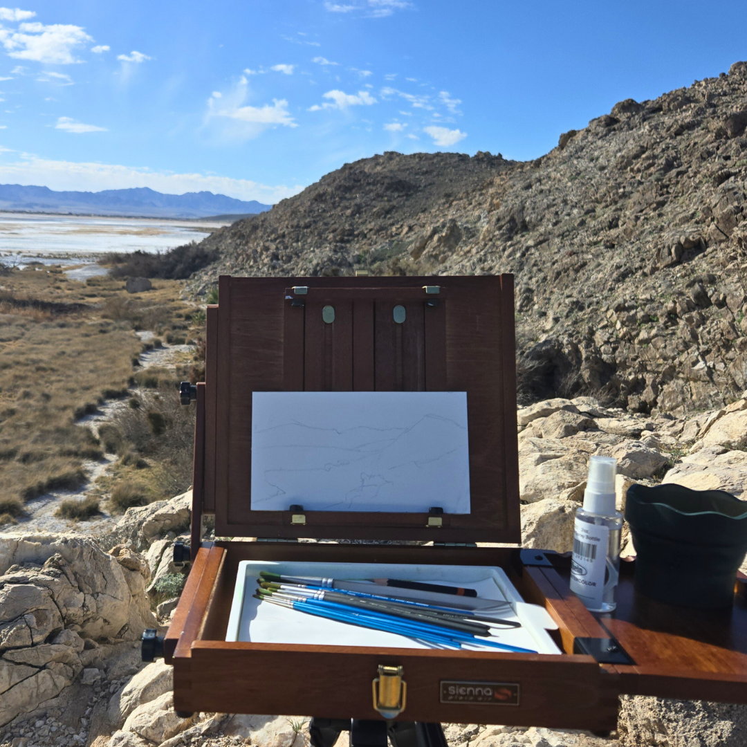 Plein air painting setup on rocky desert terrain, with an open wooden paint box, brushes, and a blank sketch, mountains and open sky in the background.