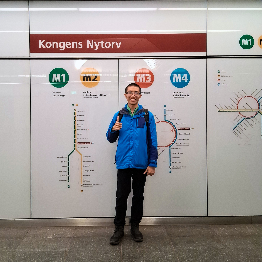 Jefferson stands smiling in a blue jacket, giving a thumbs-up in front of a wall-sized Copenhagen Metro map under a “Kongens Nytorv” station sign.