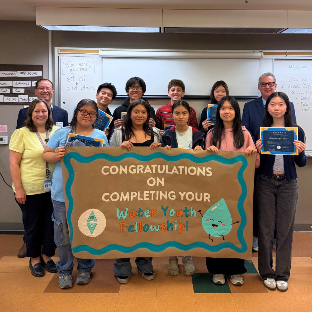 A group of students and teachers pose in a classroom holding certificates and a large sign that reads “Congratulations on completing your Water Youth Fellowship!”
