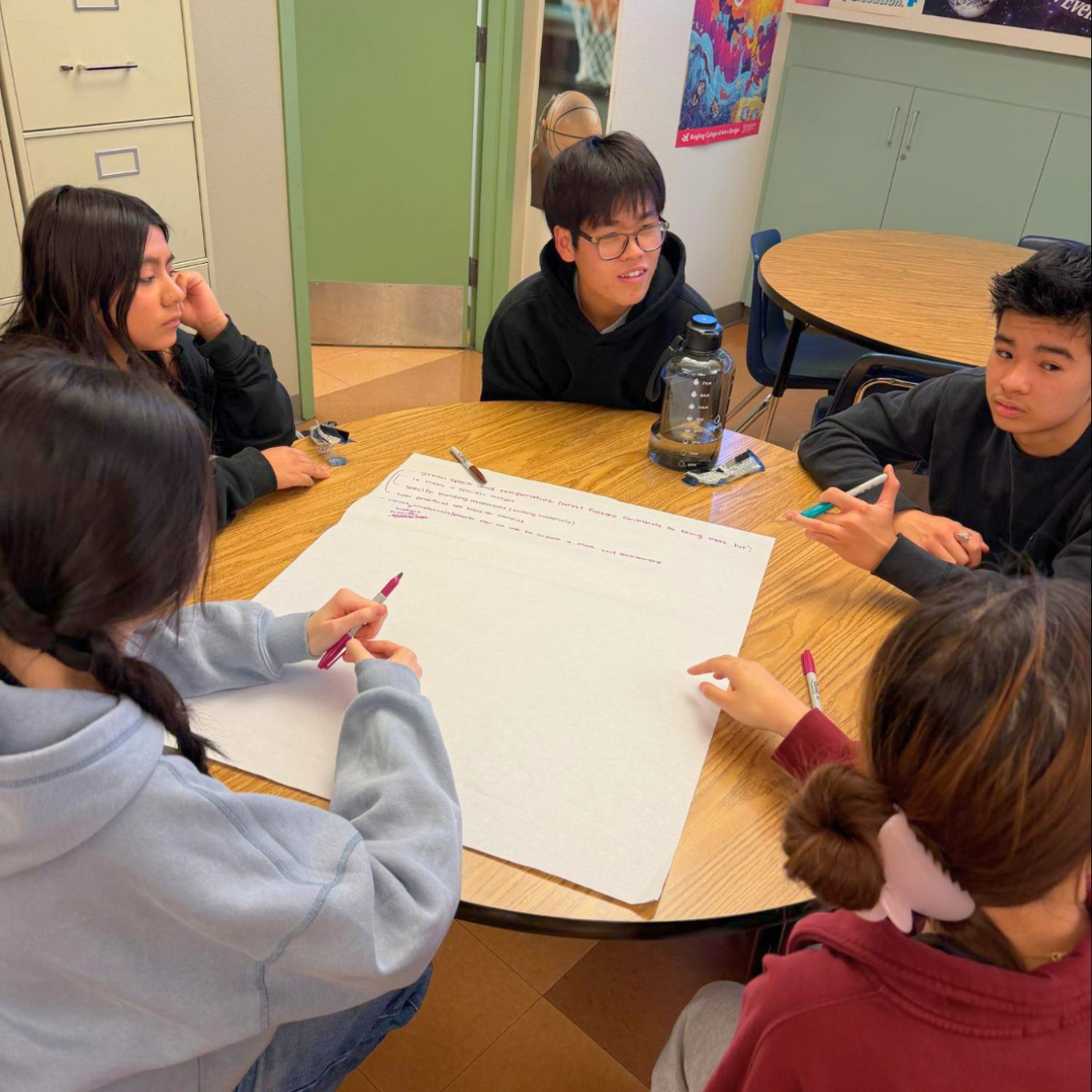 Students gather around a table with a large poster sheet during a group discussion.