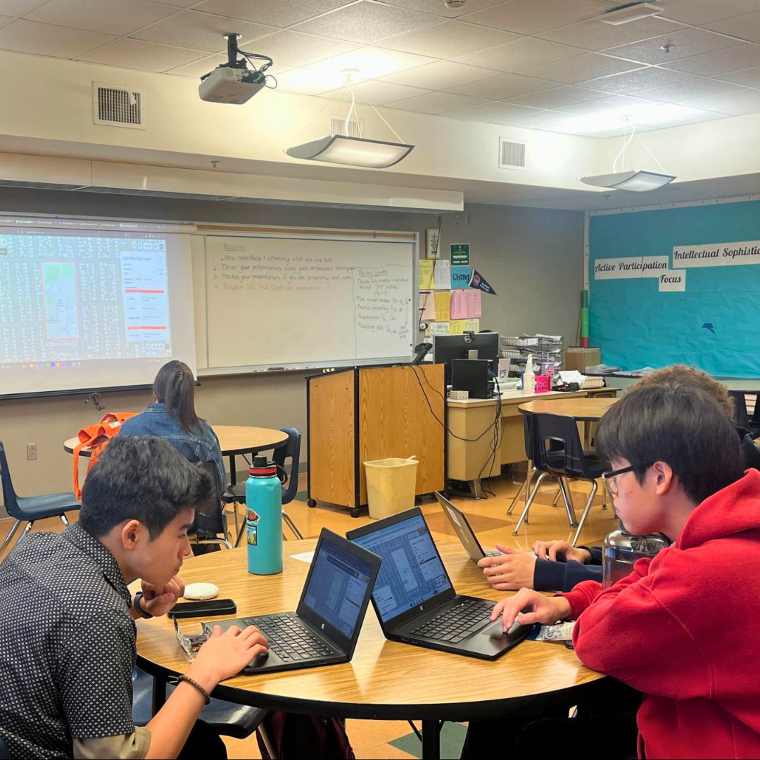 Students use laptops in a classroom while a presentation is projected at the front.