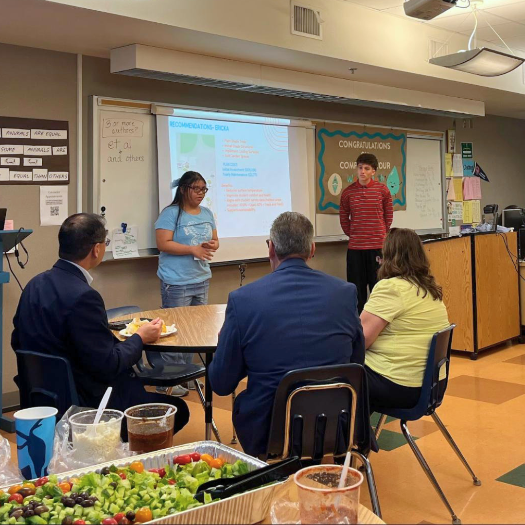 Two students speak in front of a projected presentation while adults listen.