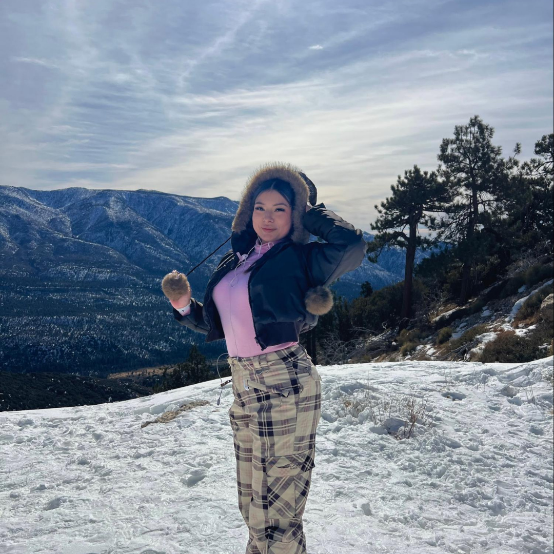 Jaylene poses in a snowy mountain landscape wearing winter clothes, with trees and mountain ridges in the background.