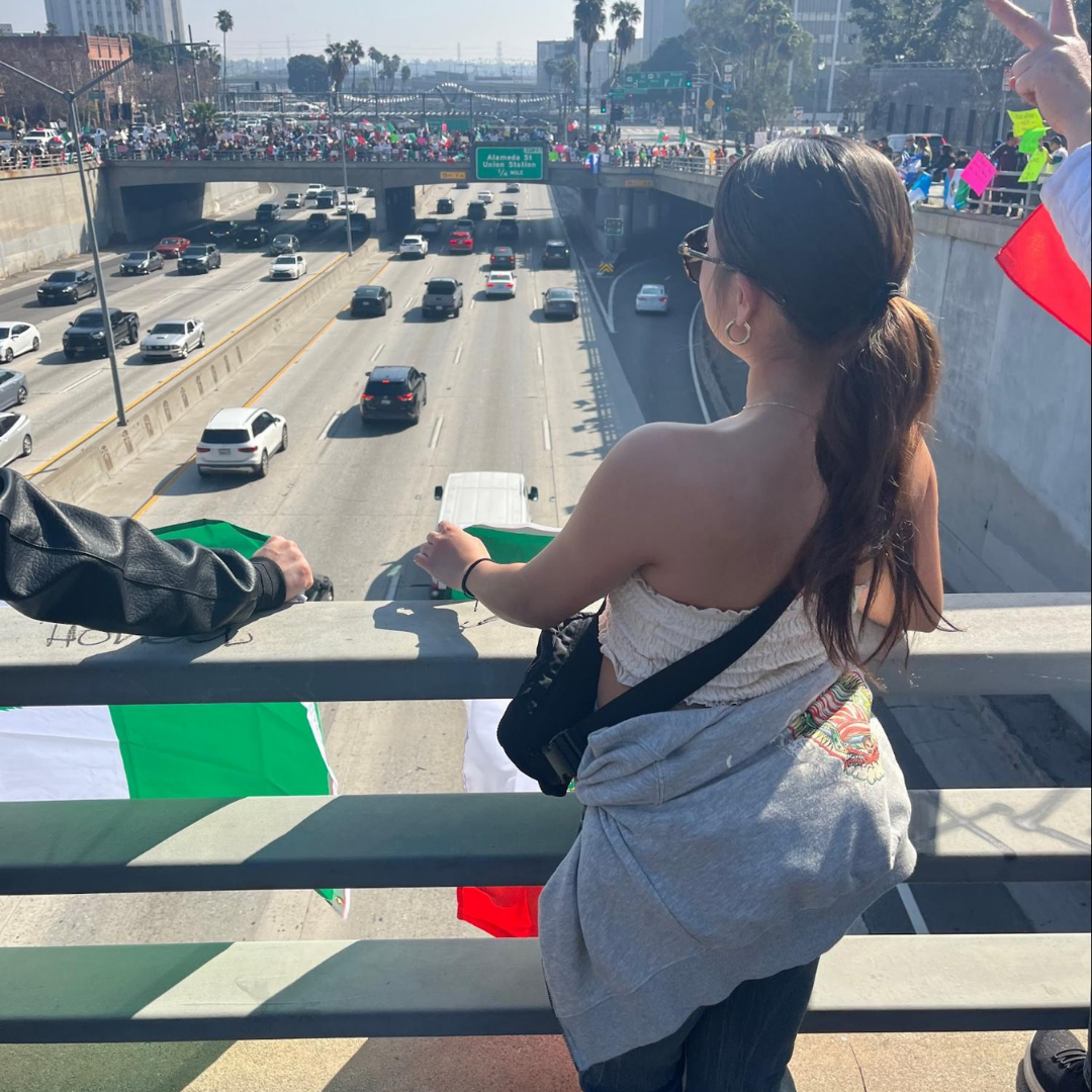 Jaylene looks out from an overpass above a freeway while holding a flag, with a crowd gathered on the bridge ahead.