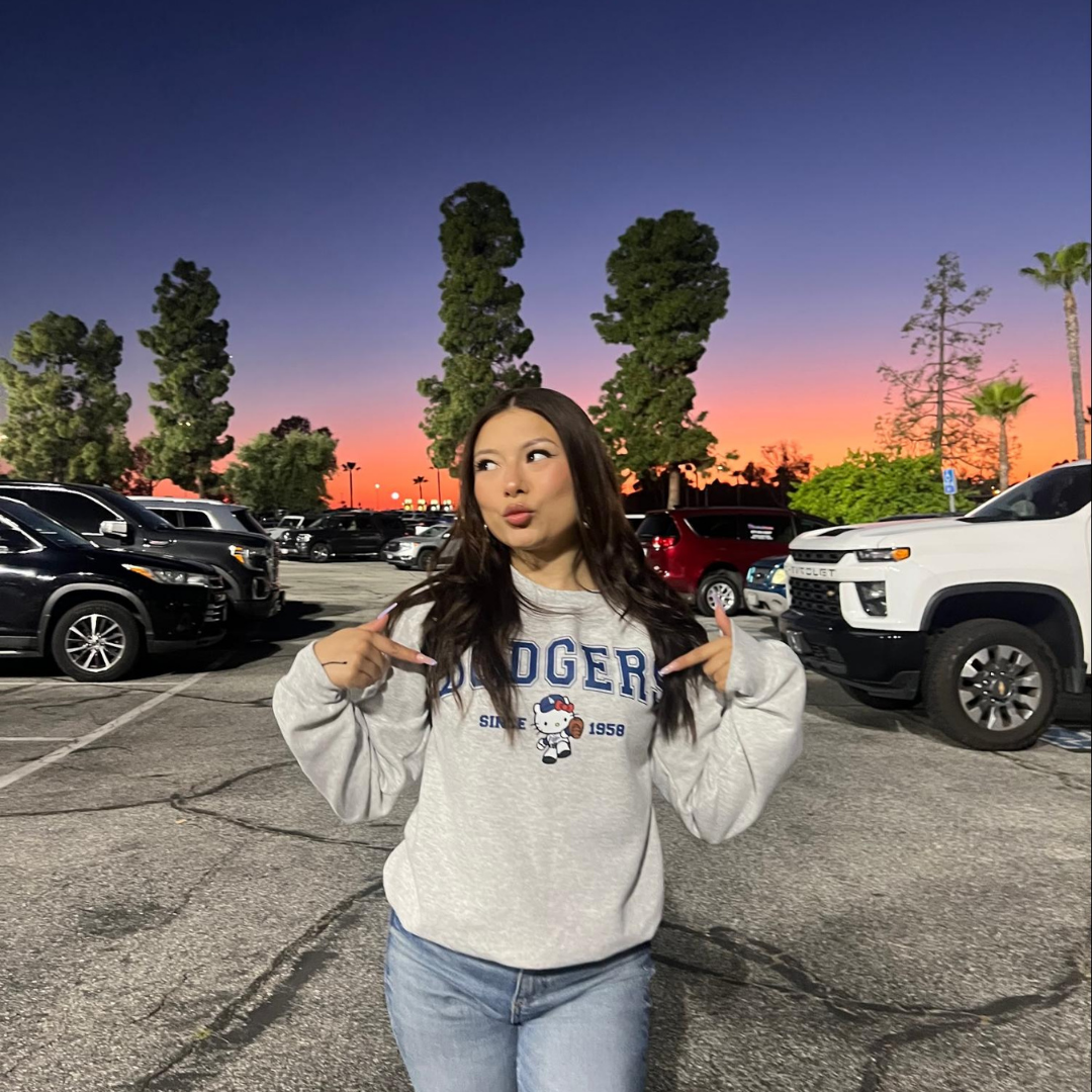 Jaylene stands in a parking lot at sunset wearing a gray Dodgers sweatshirt and jeans, pointing toward her shirt, with colorful evening sky in the background.