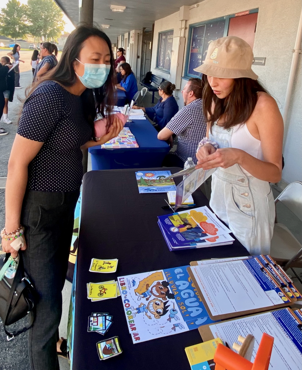 A woman wearing a mask engages with a representative at an information table during a community event. The table is covered with colorful educational materials, including flyers, brochures, and a children’s coloring book about water conservation. The representative, wearing a beige bucket hat and casual clothing, is explaining one of the pamphlets. Several other tables with seated individuals are visible in the background, and the setting appears to be outdoors at a school or community center.