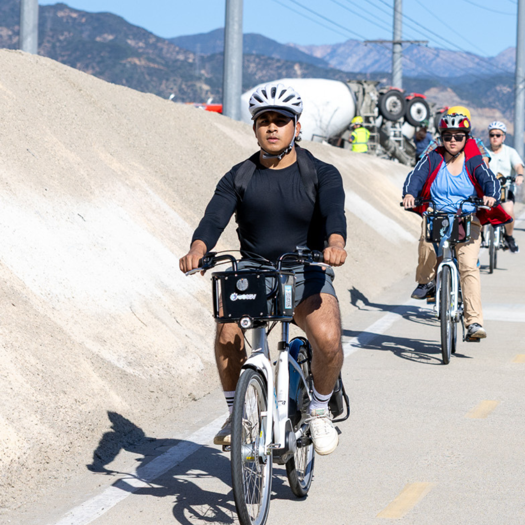 A group of people riding e-bikes along a paved river path with mountains in the background.